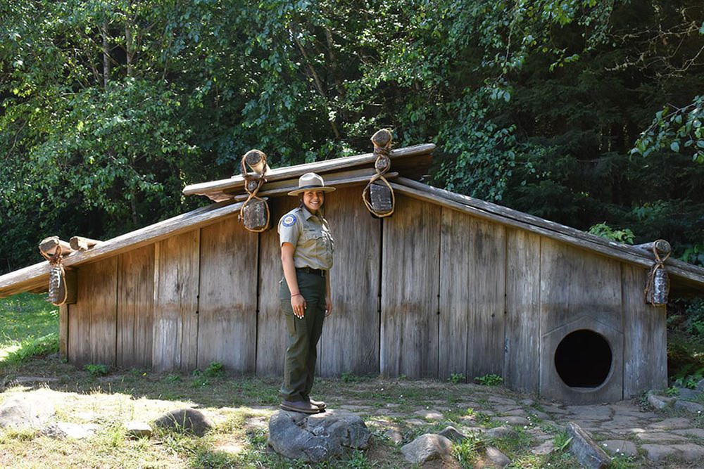 A park ranger stands in front of a wooden hut and smiles at the viewer