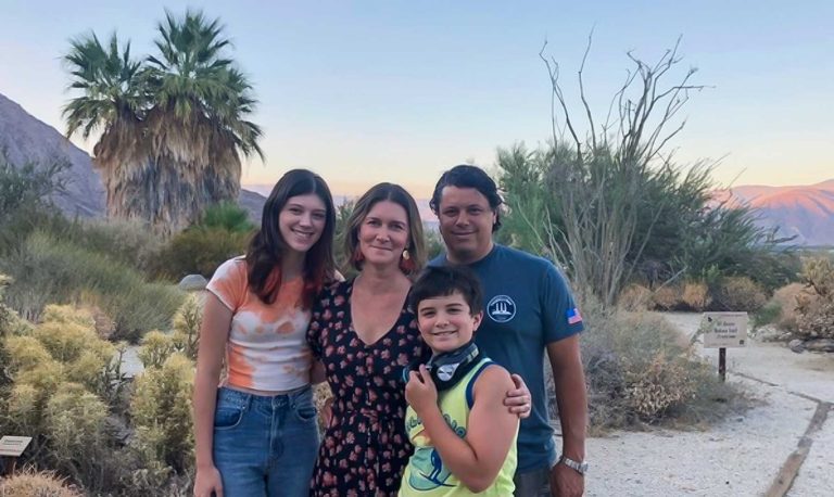 Family of four smiling in a desert park with palm trees and mountains in the background.